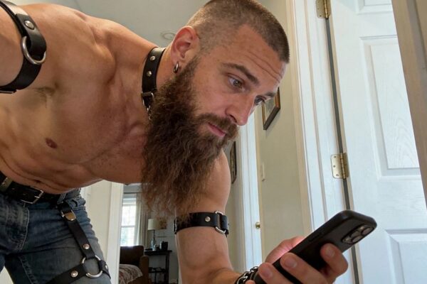Muscular man with a long brown beard wearing black leather straps, a collar, and blue jeans while looking at his phone