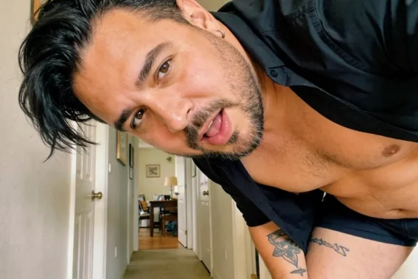Stocky dark-haired man with trimmed facial hair leaning toward the camera in an open black shirt and black shorts